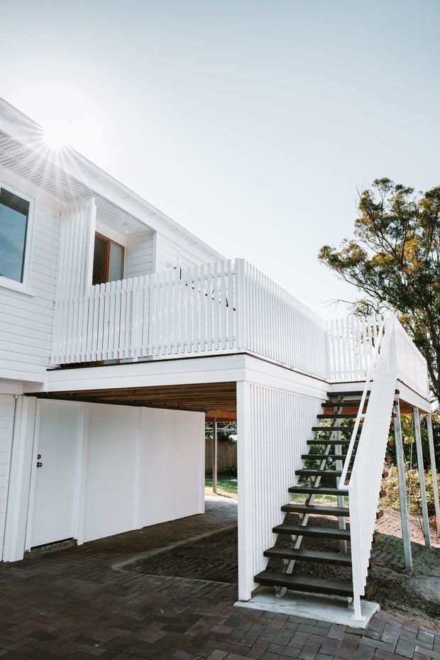 Exterior view of a raised white weatherboard house with a covered undercroft and paved parking or access area. A white-railed upper balcony or deck wraps around the building, with an external open-riser staircase leading up to it. Vertical white balustrade detailing and timber-framed windows define the facade.