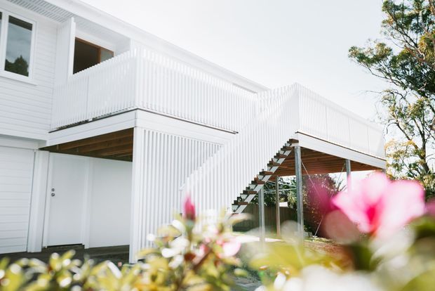 Exterior view of a bright white, two-storey residential building with a raised timber deck and balcony, white vertical balustrades, an external staircase, white weatherboard-style cladding, and exposed timber soffits. Blurred flowering planting occupies the foreground.