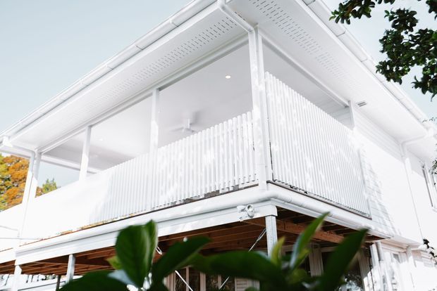 Upward-looking exterior view of a bright white, elevated two-storey residential building with a covered balcony, white vertical balustrade screening, exposed timber soffit beneath the upper level, and white guttering and downpipes. Tropical foliage partially frames the foreground.