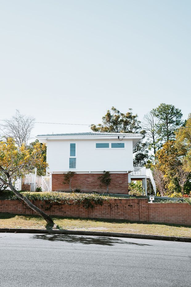 Street-facing exterior view of a raised suburban house with white horizontal cladding over a red brick lower level, a low-pitched corrugated metal roof, simple rectangular windows, and an external stair leading to an elevated deck. The property is enclosed by a brick boundary wall and timber fencing with mature trees around it.