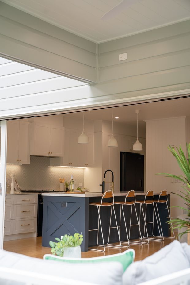 A bright, refined open-plan kitchen viewed from an adjacent living area. The kitchen features pale cabinetry, a light stone benchtop, a dark blue-grey island with framed detailing, black appliances and tapware, a subtle herringbone splashback, pendant lights, and a row of contemporary white-framed bar stools. The room opens to a covered glazed area or skylit opening, with a sofa and plants partially visible in the foreground.
