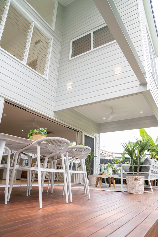Bright covered outdoor entertaining deck adjoining an open-plan interior, with a dining table and woven chairs, potted tropical plants, white horizontal weatherboard cladding, louvred windows, recessed lighting and a ceiling fan. The elevated camera angle emphasizes the timber decking and sheltered indoor-outdoor connection.