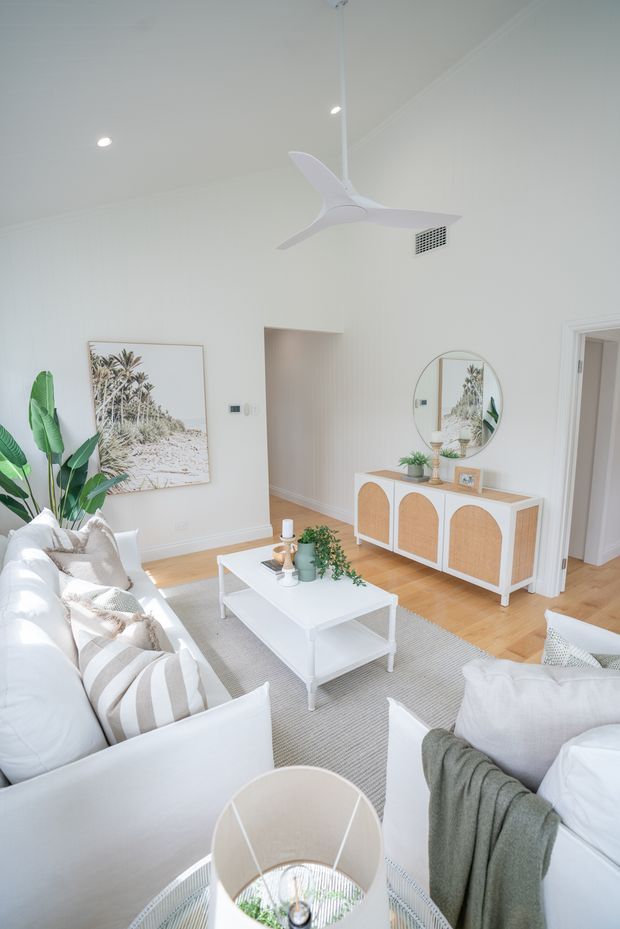 Bright, airy living room with white walls and trim, light timber flooring, white sofas, a pale striped rug, and a white coffee table. A cane-front sideboard with a round mirror, indoor plants, framed coastal artwork, and a sculptural white ceiling fan create a relaxed contemporary coastal aesthetic.