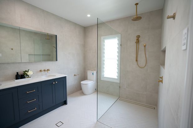 A bright, contemporary bathroom with a dark navy-blue vanity, pale stone-look wall tiles, white herringbone floor tiles, a wall-mounted toilet, and a frameless glass walk-in shower. Brass fixtures, including the shower fittings, taps, handles, and wall light, provide warm accents. A mirrored cabinet spans the vanity wall, and a louvred window brings in natural light.