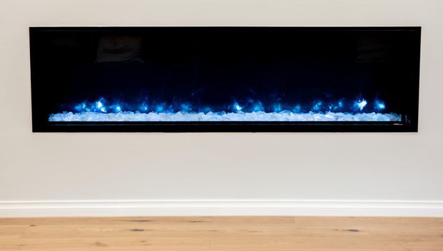 A minimalist interior wall featuring a long, recessed black electric fireplace with blue flame-like lighting and white ember stones. The fireplace is set into a plain light-coloured wall above simple white skirting, with pale timber flooring below.