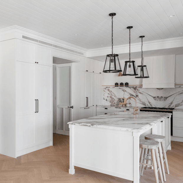 A bright, refined white kitchen with extensive built-in cabinetry, a large marble island and matching marble splashback, brass tapware, black range cooker, and three suspended black metal pendant lights. The warm herringbone timber floor and pale wood stools add contrast to the predominantly white scheme.