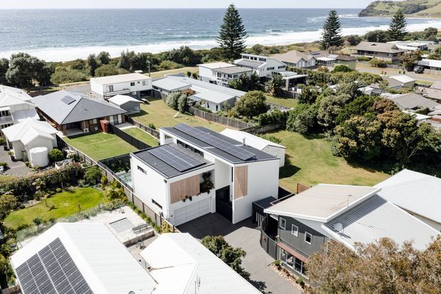 Aerial view of a suburban coastal neighbourhood with a prominent modern flat-roofed white house featuring extensive rooftop solar panels, dark-framed glazing, and timber-toned vertical screening. Surrounding homes, landscaped gardens, streets, and the ocean with a rocky headland are visible in the background.