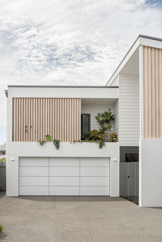 Front exterior view of a contemporary multi-level residential home with a sharply pitched gable roof, white horizontal cladding, clean geometric forms, a large segmented garage door, and a covered upper-level balcony or entry screened by vertical timber-toned battens. Integrated planter boxes soften the minimalist facade.