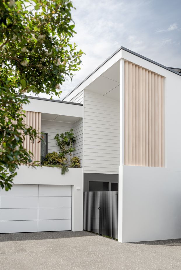A contemporary residential facade viewed from the driveway, featuring crisp white rendered walls, horizontal weatherboard cladding, a sharply pitched roof form, vertical pale timber or slatted screening, a white sectional garage door, and a dark slatted pedestrian gate. A planted balcony or entry courtyard is partially screened by mature foliage.