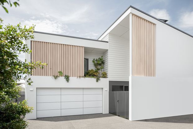 Contemporary two-storey residential facade with crisp white walls, a flat and skillion roof composition, integrated garage, and tall recessed entry volume. Vertical timber-look screening adds warmth and privacy, with balcony planter greenery and a dark driveway completing the minimal exterior.
