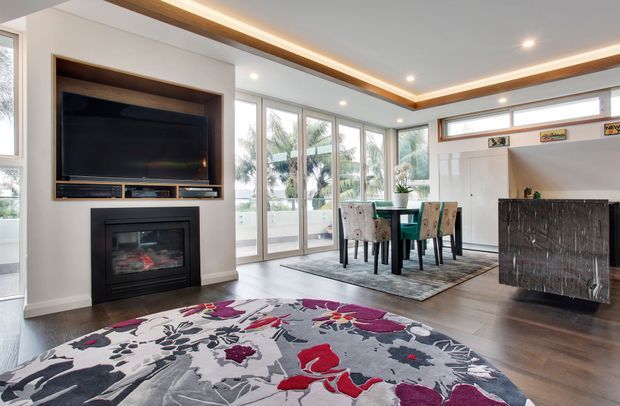 Bright contemporary open-plan living and dining area with a built-in dark fireplace and television wall, timber-trimmed recessed ceiling detail, large corner glazing, a dining table with upholstered chairs, and a striking floral area rug. A compact dark bar or storage cabinet sits to the right.