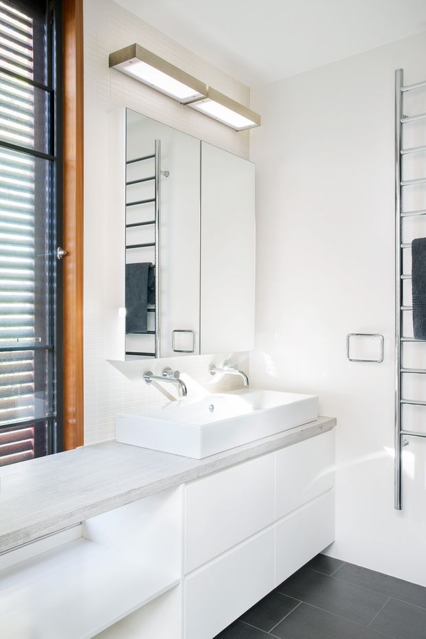 Bright contemporary bathroom with a white wall-mounted vanity, integrated rectangular basin, mirrored medicine cabinet, and chrome fixtures. A vertical chrome heated towel rail is mounted on the right wall, while a timber-framed louvred window provides daylight on the left. The room has a dark grey tiled floor, white tiled walls, and a pale grey benchtop.
