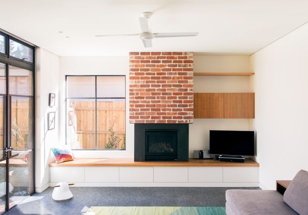 Bright contemporary living room with a white ceiling and walls, large black-framed glazing, a red exposed-brick fireplace chimney, and a built-in timber and white cabinetry wall. The fireplace, television, low joinery, grey carpet, sectional sofa, and ceiling fan create a clean, warm, functional interior.