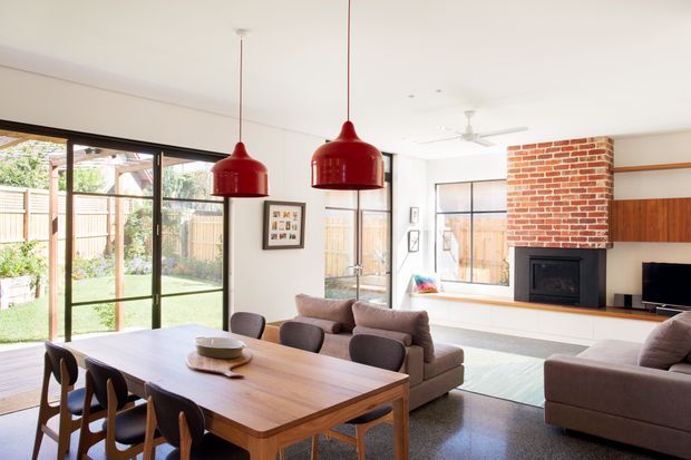 Bright open-plan living and dining area with a large timber dining table, upholstered dining chairs, sofas, a brick fireplace surround, timber wall panelling, broad windows and sliding doors opening to a landscaped backyard. Two oversized red pendant lights provide bold focal points over the dining table.