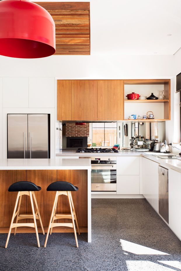 Bright contemporary kitchen with white cabinetry, warm timber island and overhead joinery, a white benchtop, stainless-steel appliances, open shelving, and a red pendant light. Two bar stools sit at the island, while a brick fireplace is visible beyond the cooking area. The floor is a speckled polished concrete or terrazzo-like finish, and daylight fills the space.