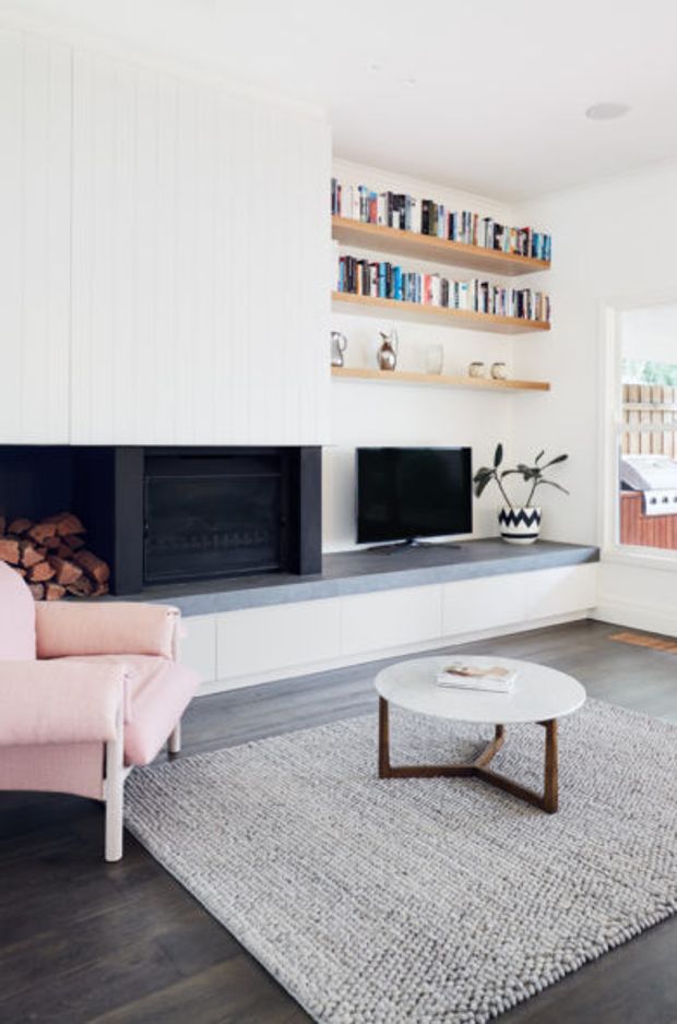 Bright contemporary living room with a white vertical-panelled built-in media and fireplace wall. The fireplace has dark surrounds and integrated timber log storage, while open timber shelves display books and ceramics. A low white built-in entertainment cabinet sits beneath a wall-mounted television. The room includes a pale pink armchair, round coffee table, textured light rug, dark timber flooring, and a large window opening to an outdoor deck.