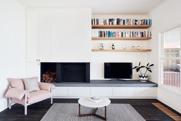 Bright, compact living room with a built-in white wall unit incorporating a black fireplace, firewood storage, open timber display shelves, and low media cabinetry. A television, potted plant, pale pink armchair, round coffee table, and textured rug sit over dark timber flooring. White walls, ceiling mouldings, and large windows create an airy contemporary interior.