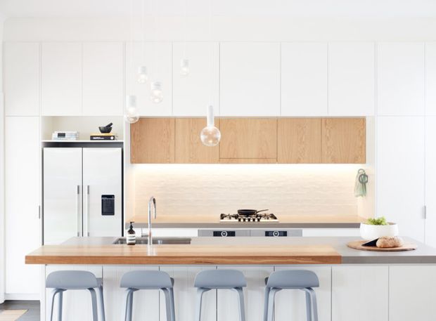 Bright, clean contemporary kitchen with white full-height cabinetry, a timber island benchtop, integrated appliances, pale timber overhead cabinets, a softly illuminated splashback, pendant lights, and four stools at the island.