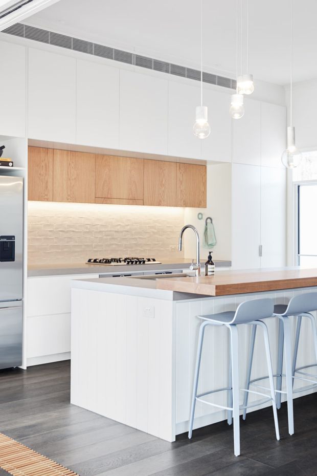 Bright contemporary kitchen with a large central island, white full-height cabinetry, warm timber overhead cupboards and island benchtop, pale textured brick splashback, dark timber flooring, and pale blue bar stools. Globe pendant lights hang above the island, with integrated appliances and a gas cooktop along the rear wall.
