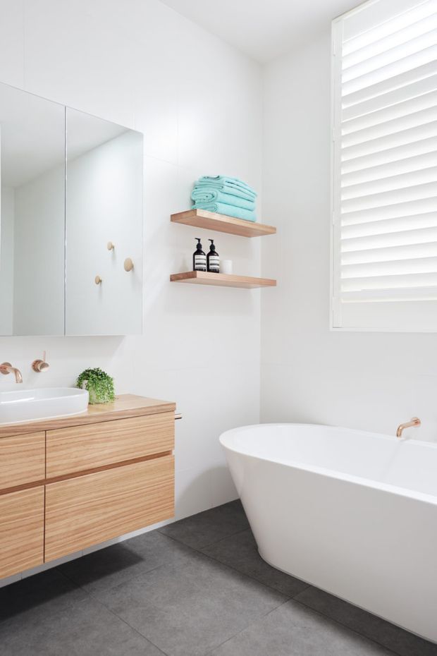 Bright contemporary bathroom with white walls and a large white plantation-shutter window. A floating timber vanity with integrated white basin sits beneath mirrored cabinetry, complemented by open timber shelves, black-and-white dispensers, and folded aqua towels. A freestanding white bathtub with a brass tap occupies the right side, while large dark grey floor tiles provide contrast.
