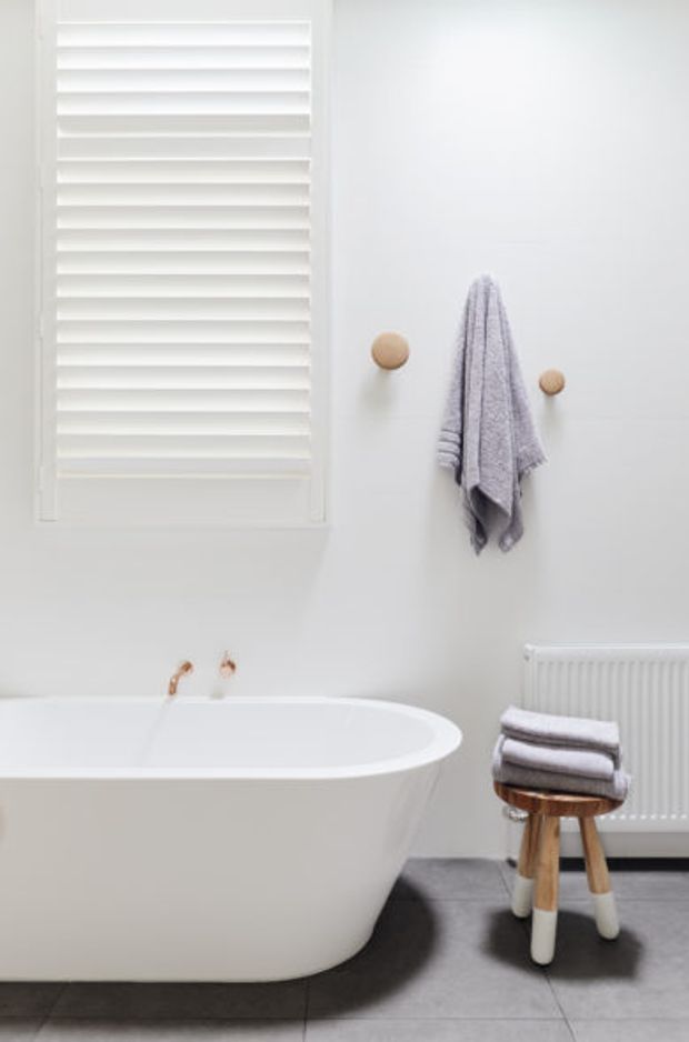 Bright, minimalist bathroom featuring a freestanding white bathtub, white plantation shutters, a wall-mounted towel with round wooden hooks, a white radiator, and folded towels on a small wooden stool. The floor is finished in dark grey tiles with a clean, restrained palette.