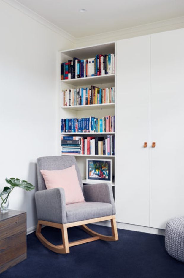 A compact, thoughtfully designed reading nook with a grey upholstered rocking armchair, pale pink cushion, dark navy carpet, and a built-in white bookcase and storage cabinetry. The white joinery incorporates open shelving filled with books, enclosed cupboards with small timber knobs, and a small framed artwork. White walls and decorative cornices create a clean, refined interior, while a timber side table and indoor plant add warmth.