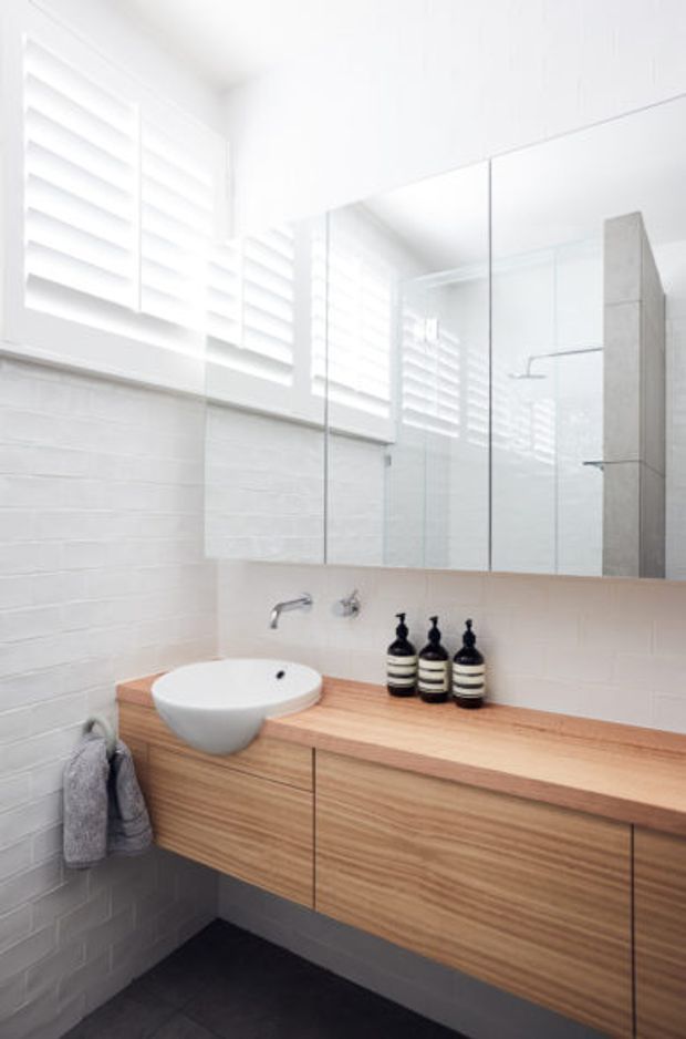 Bright, compact contemporary bathroom with a wall-mounted timber vanity, white vessel basin, large mirrored cabinet, white textured wall tiles, dark floor tiles, and a frameless glass shower enclosure. Plantation shutters provide filtered natural light.