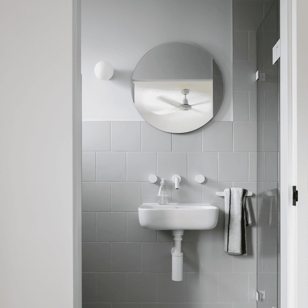 A compact, bright bathroom viewed through an open doorway. The room features pale grey rectangular wall tiles, a wall-mounted white basin with exposed plumbing, minimalist white tapware, a round mirror, a small wall light, and a towel hanging beside a glass shower screen. The mirror reflects a ceiling fan and adds depth to the small space.