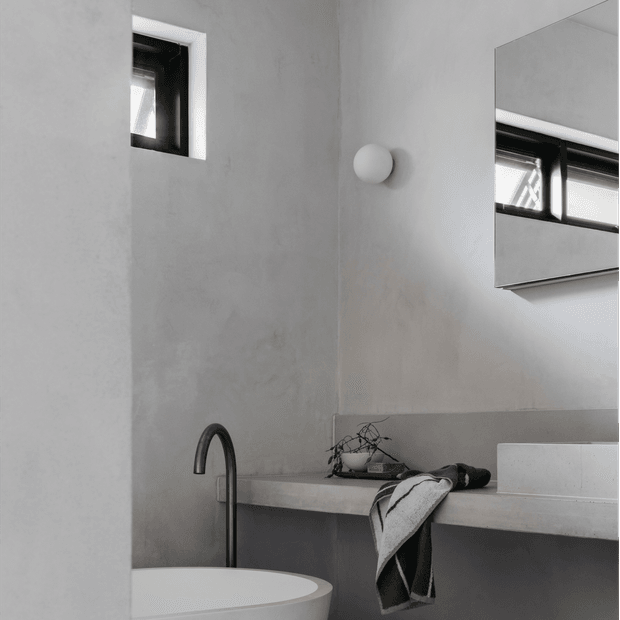 Minimalist monochromatic bathroom interior featuring a freestanding round basin, slender dark curved tap, pale stone or concrete-look vanity, large wall mirror, small high-set black-framed window, and a spherical wall light. The space has smooth light grey walls and a restrained contemporary fit-out.