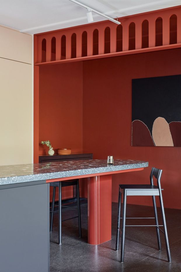 A richly coloured contemporary kitchen and dining area dominated by deep terracotta-red built-in joinery and walls. A large stone island with a speckled polished surface extends across the foreground, supported by a rounded red column and paired with dark bar stools. The rear wall features integrated cabinetry with repeated tall arched niches, a low dark sideboard, and abstract artwork. A pale contrasting wall, dark flooring, and ceiling-mounted track lighting complete the space.