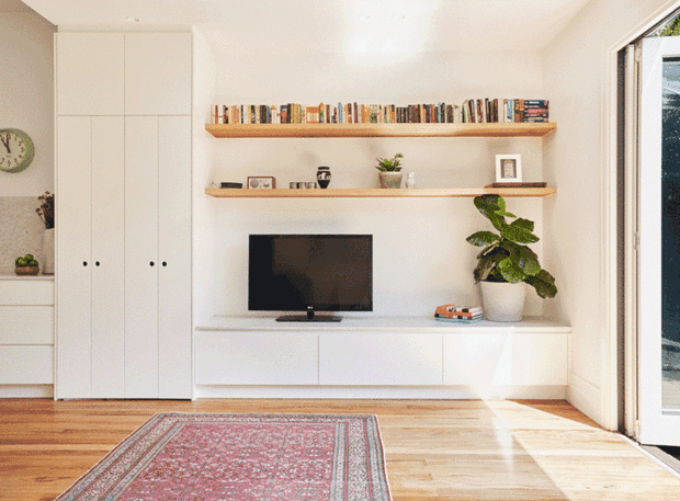 Bright, compact living room with white built-in cabinetry and low media storage, open timber display shelves filled with books and small decor, a television, indoor plant, timber flooring, and a patterned area rug. Large glazed opening on the right brings in strong natural light, creating a clean, warm, contemporary interior.