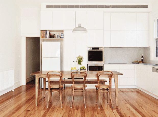 Bright, compact open-plan kitchen and dining area with full-height white flat-panel cabinetry, integrated appliances, pale stone benchtops and splashback, timber flooring, and a rustic timber dining table with four bentwood chairs. A simple pendant light hangs above the table, while open shelving and a concealed refrigerator add practical storage. The space has a clean, warm, understated contemporary character.