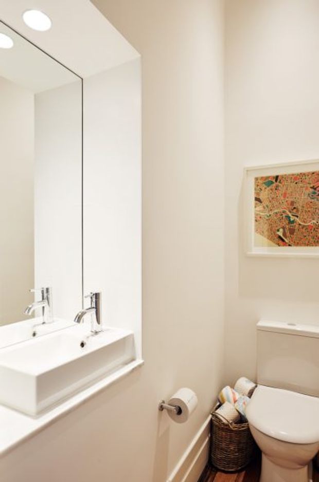 Compact bright bathroom with a white pedestal-style basin and chrome tapware beneath a large frameless wall mirror. A white toilet sits beside the vanity, with a woven basket and framed artwork adding small decorative details. The walls and fixtures are predominantly white and warm off-white, with recessed ceiling lighting and a narrow timber-toned floor visible at the base.