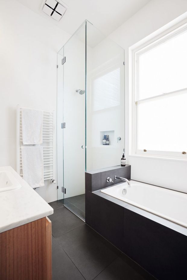 Bright, contemporary bathroom featuring a full-width freestanding-style bathtub, dark built-in vanity with white basin and chrome tapware, frameless glass shower enclosure, dark floor tiles, white walls, a heated towel rail, and a large white-framed window. A timber vanity element is visible at the left edge.