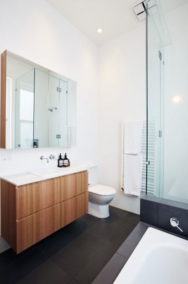 Bright contemporary bathroom with white walls, a wall-mounted timber vanity with integrated white benchtop, large mirror, wall-hung toilet, dark floor tiles, glass shower enclosure, heated towel rail, and a partially visible freestanding bathtub. The space is clean and compact with strong black-and-white contrast and warm timber joinery.