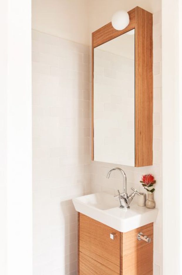 Compact, bright bathroom vanity area featuring a wall-mounted timber vanity with a white integrated basin, chrome mixer tap, matching timber-framed mirrored cabinet, spherical wall light, and white tiled walls. Small decorative vase and flowers sit on the basin edge.
