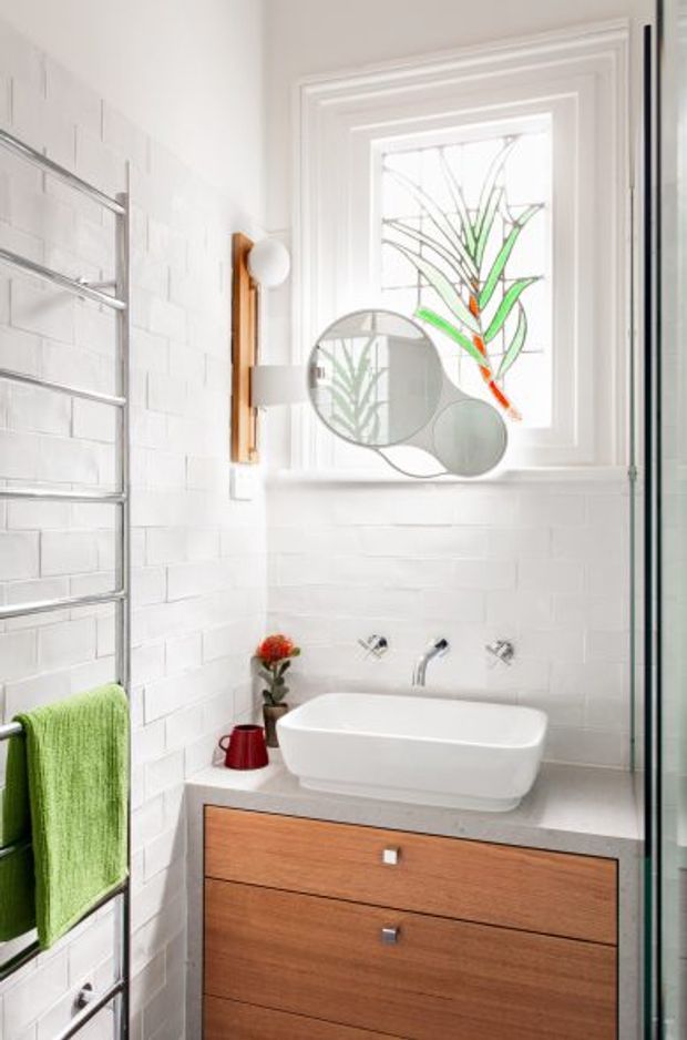 Bright compact bathroom featuring white subway-tile walls, a wall-mounted timber vanity with a white countertop and vessel basin, chrome wall-mounted taps, a heated towel rail with a green towel, and a decorative frosted stained-glass window. A round multi-panel mirror and simple wall light add visual interest.