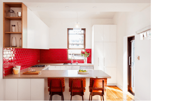 A bright, compact contemporary kitchen with white flat-panel cabinetry, a large pale island with three timber bar stools, and a vivid glossy red subway-tile splashback. Open timber shelving with simple kitchenware sits at the left, while warm timber flooring and a glazed rear door add contrast. The room receives strong natural light and includes a central pendant light.