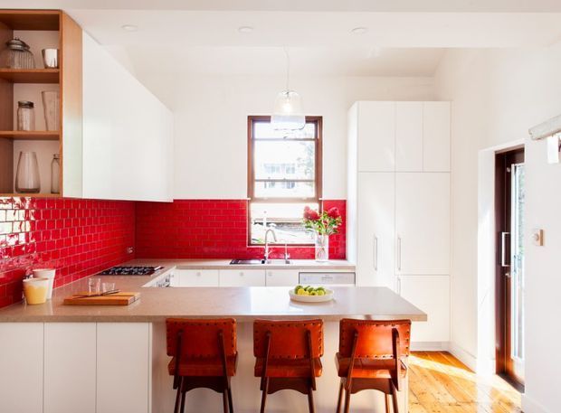 Bright contemporary kitchen with white flat-panel cabinetry, a large central island with three leather bar stools, a red glossy subway-tile splashback, pale stone benchtops, timber flooring, open timber shelving, and a window above the sink. The room has abundant natural light and a warm, high-contrast palette.