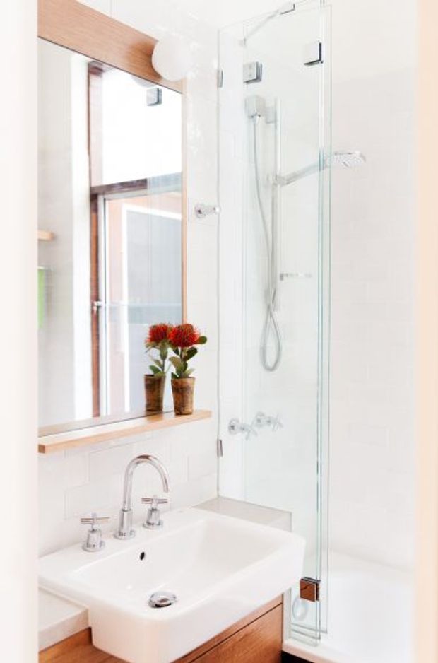 Bright compact bathroom with a white wall-mounted basin, chrome tapware, timber vanity cabinetry and timber-framed mirrored storage. A clear glass shower enclosure with a handheld shower and wall-mounted showerhead occupies the right side. White tiled walls and a small timber-framed window ledge with potted plants create a clean, light-filled interior.