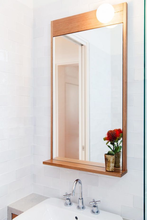 Bright bathroom vanity area featuring a large rectangular mirror with a warm timber frame and integrated timber shelf, mounted over a white ceramic basin with chrome tapware. The surrounding walls are clad in glossy white rectangular tiles, and a small potted flowering plant adds colour beside the mirror.
