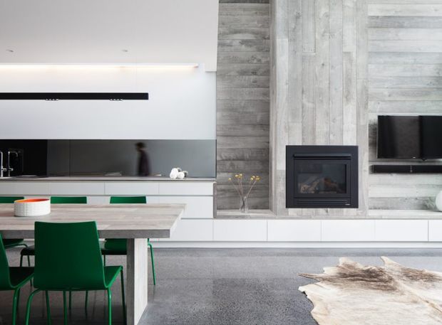 Open-plan contemporary living and dining area with a sleek white kitchen, long island bench, vivid green dining chairs, and a black reflective splashback. A grey timber-look feature wall incorporates a built-in fireplace and wall-mounted television, while a pale hide rug sits over grey carpet.