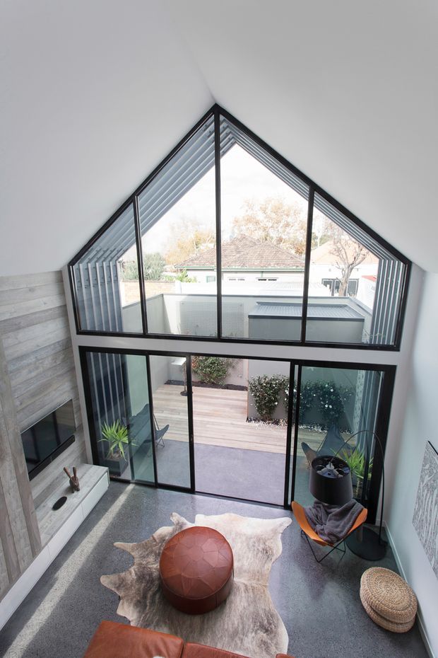 A dramatic double-height living room with a steeply angled glazed gable wall and black-framed floor-to-ceiling windows overlooking a courtyard. The room features polished concrete flooring, a grey stone or concrete fireplace wall with built-in television, a low leather sofa, a sculptural round leather ottoman, a hide rug, indoor plants, and an outdoor timber deck visible beyond the glazing. The composition emphasizes the tall glazed void, abundant natural light, and strong indoor-outdoor connection.