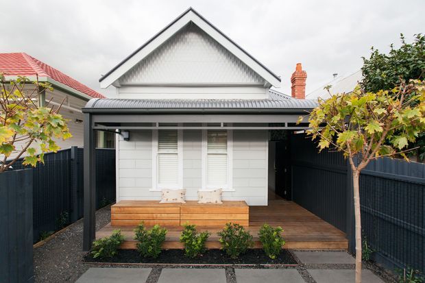 Front exterior view of a compact single-storey gabled home with a white weatherboard facade, corrugated metal verandah roof, covered timber porch, built-in timber bench seating, dark fencing, and a landscaped entry garden. The traditional gable form is paired with a contemporary front addition and crisp black-and-white detailing.