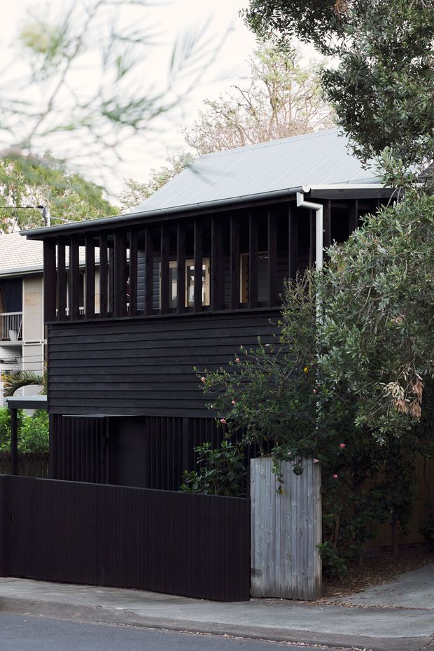 Street-facing exterior of a compact dark-clad dwelling, with a pitched light-metal roof, deep vertical screening around the upper-level glazing, horizontal timber-style wall cladding, and a dark privacy fence. Mature vegetation partially frames the facade, while a small adjoining structure is visible at left.