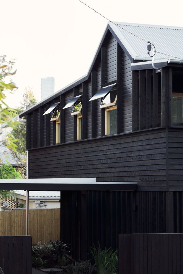 Exterior view of a compact dark-clad multi-level building in an urban residential setting. The facade features black timber weatherboard cladding, a steep gabled corrugated metal roof, tall narrow windows with projecting metal awnings, and a covered lower-level carport or sheltered entry. Fencing and planting are visible at ground level, with neighbouring urban buildings in the background.