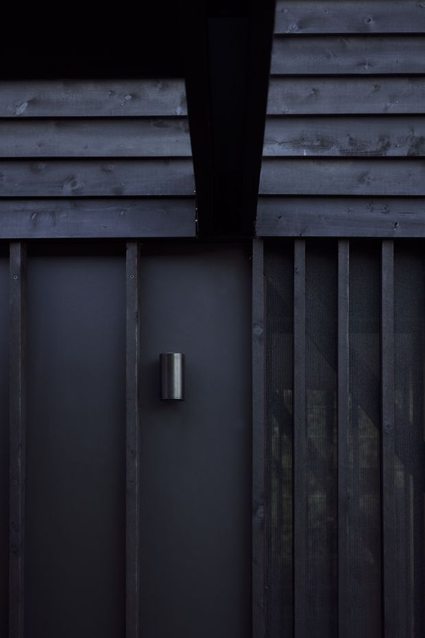 Close-up detail of a dark, moody building facade. Weathered horizontal timber cladding sits above vertical timber battens, with a dark recessed door or wall panel and a small cylindrical metal wall light. Translucent or screened glazing is visible behind the battens on the right.
