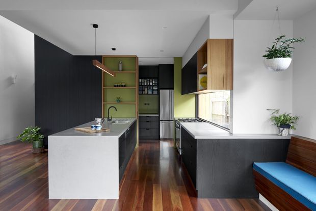A contemporary galley-style kitchen with a central white island, dark charcoal cabinetry, timber open shelving, and a pale green splashback or rear wall. The room features polished dark timber flooring, white stone benchtops, integrated appliances, pendant lighting, and a built-in timber bench seat with blue upholstery. Indoor plants add a natural accent.