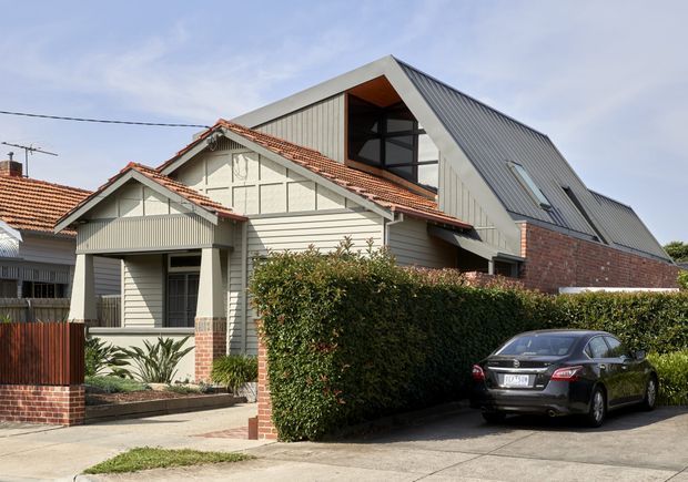 Exterior view of a residential house combining an older single-storey home with a prominent contemporary rear or upper extension. The building features cream weatherboard cladding, red brickwork, decorative gable detailing, terracotta tiled roofing on the original section, and a steep grey metal gable roof with large dark-framed glazing on the addition. A hedge-lined driveway and front garden are visible.