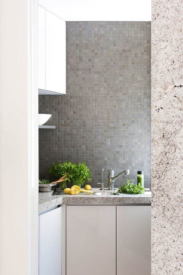 A compact contemporary kitchen viewed through a doorway, featuring white and pale grey cabinetry, a speckled stone benchtop, stainless-steel mixer tap and sink, and a full-height grey mosaic tile splashback. Fresh produce and herbs sit on the benchtop, adding natural green and yellow accents.